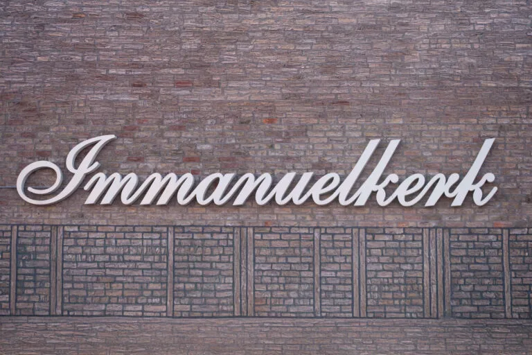 Wit sierlijk gevelopschrift ‘Immanuelkerk’ op een muur van rode en grijze bakstenen.