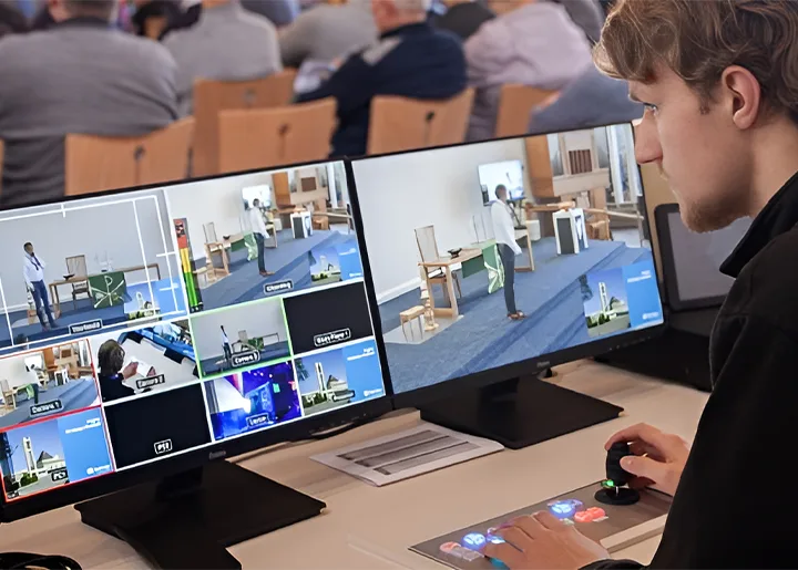 Technicus bedient een livestreamregie met meerdere schermen waarop beelden van een kerkdienst worden weergegeven; op de achtergrond zit het publiek in houten stoelen.