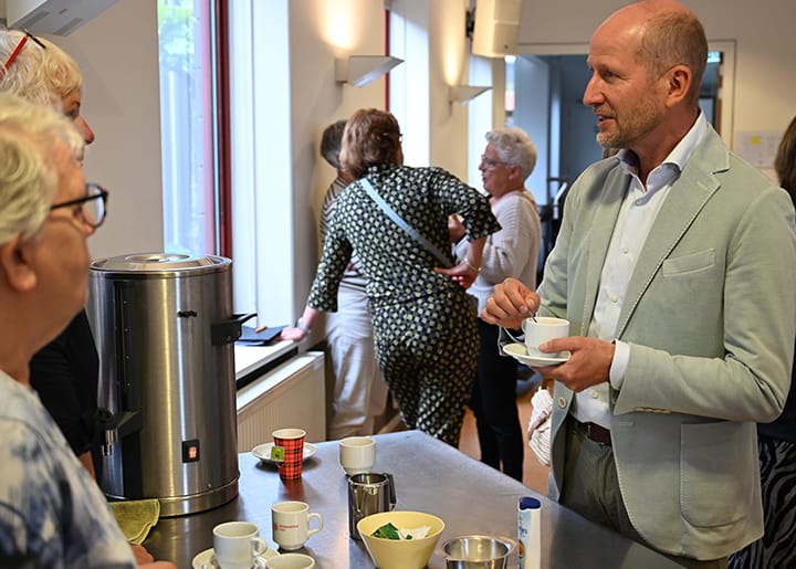 Man in licht pak in gesprek met anderen tijdens een koffiemoment in de kerk; op de voorgrond een tafel met kopjes, theezakjes en een koffiecontainer.