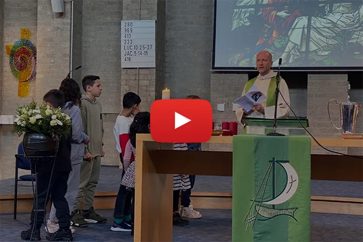 Afbeelding van een voorganger in witte liturgische kleding met groene stola die kinderen toespreekt in de kerk, met een grote rode ‘play’-knop erop om aan te geven dat het een video is.