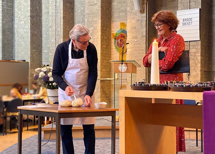 Man in schort bereidt brood voor tijdens een kerkdienst, terwijl een vrouw toekijkt en lacht.