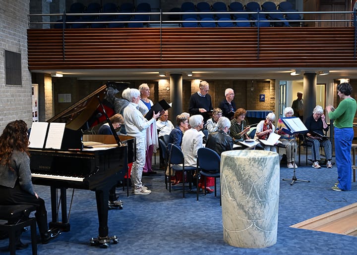 Koor repeteert in de kerk met een dirigent en pianiste, omringd door blauwe stoelen en kerkmeubilair.